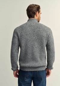Grauer Strickpullover mit hohem Kragen, strukturiertem Design und gerippten Bündchen. Tragen mit blauen Jeans, von der Rückansicht gezeigt.