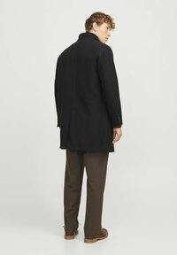 Abrigo de lana negro con cuello alto, corte recto y mangas largas. Lleva puesto unos pantalones marrones y zapatos marrones. Diseño simple y elegante.