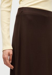 Γυναίκα που φοράει εκρού μακρυμάνικη ριμπ πλεκτή μπλούζα, μέσα σε ψηλόμεση σκούρα καφέ φούστα, με το χέρι χαλαρό στο πλάι.