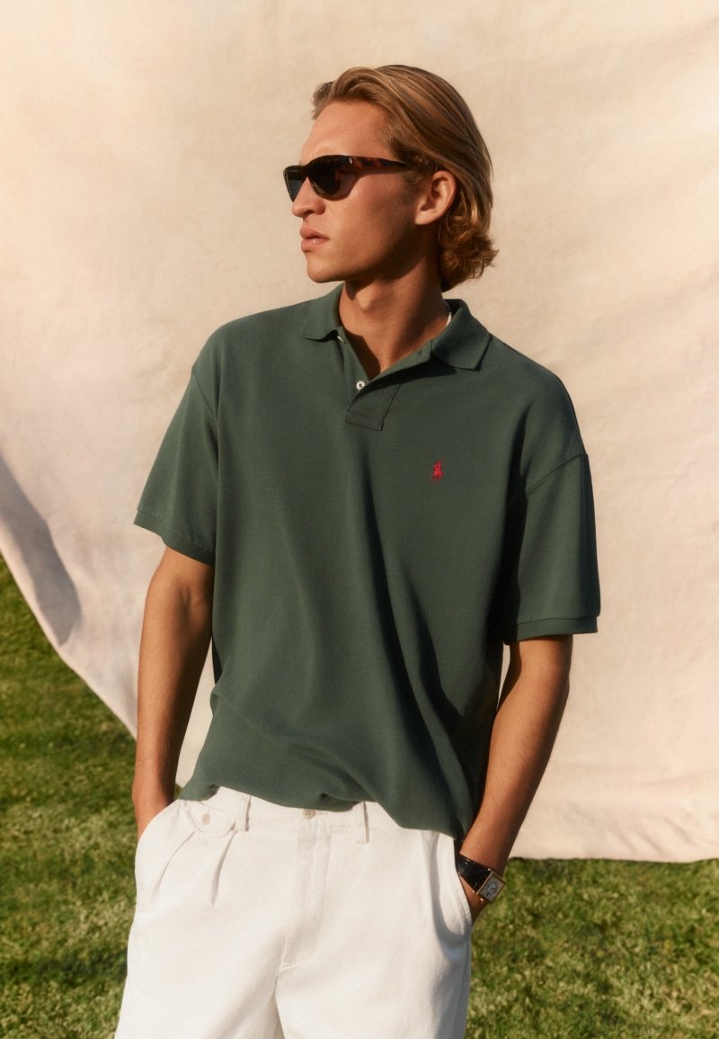 Ung mand i mørkegrøn polo-shirt og hvide bukser iført solbriller, stående med hænderne i lommerne udendørs på græs med beige baggrund.