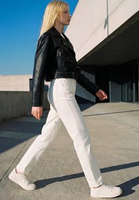 Young woman with long blond hair walking outdoors wearing a black leather jacket, white pants, and white sneakers on a concrete path.