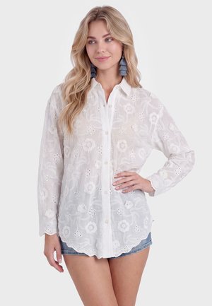 Młoda kobieta z długimi blond włosami, ubrana w białą koszulę z haftem kwiatowym i szorty z denimu, pozuje z jedną ręką na biodrze.