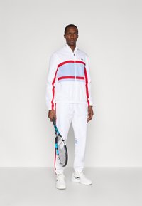 Λευκό σετ φόρμας με κόκκινες και μπλε ρίγες, μπουφάν με φερμουάρ και ψηλό γιακά, παντελόνι με ελαστική μέση και λευκά αθλητικά παπούτσια. Κρατάει μια ρακέτα τένις.