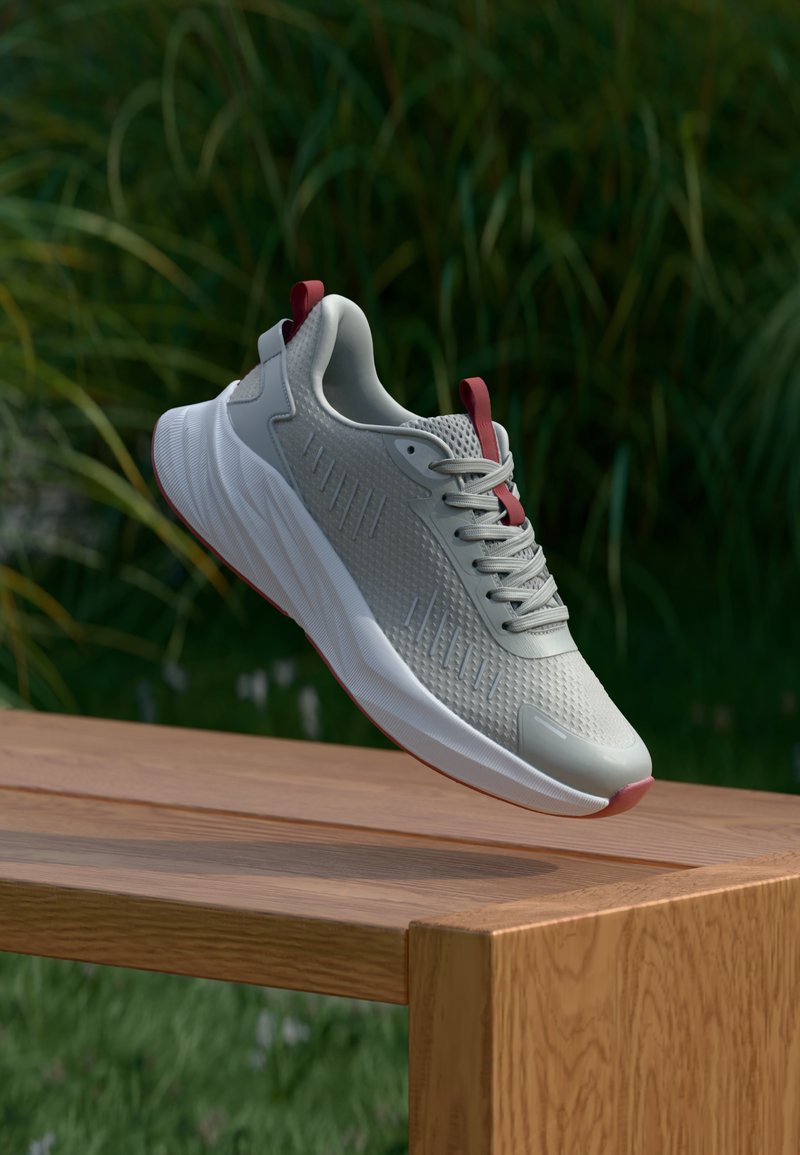 Gaiši pelēks sporta apavu pāris ar teksturētu augšdaļu, sarkaniem akcentiem, apaļu purngalu un elastīgu baltu zoli, novietots uz koka galda dārzā.