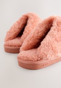 Chaussons roses duveteux avec une tige en fausse fourrure douce, une doublure rembourrée et une semelle en caoutchouc texturée. Présentent le logo "NEXT" sur le côté.