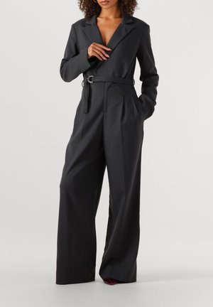 Zwarte op maat gemaakte jumpsuit met een diepe V-hals, lange mouwen, wijde pijpen en een ceintuur in de taille. Gemaakt van gestructureerde stof met zakken.