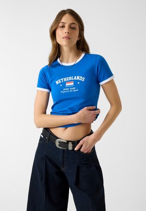 Femme portant un crop top bleu avec le texte « Netherlands » et le drapeau, associée à un jean large foncé et une ceinture noire avec une boucle argentée.