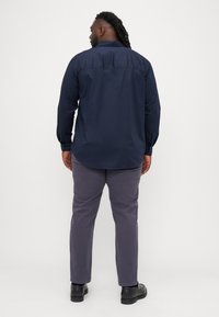 Camicia a maniche lunghe blu navy con colletto button-down, caratterizzata da un design con piega sul retro, abbinata a pantaloni grigi e scarpe nere.