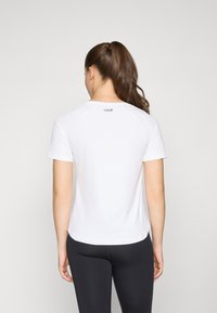 T-shirt atletico bianco a maniche corte, con texture liscia, scollatura arrotondata e logo discreto sulla parte alta della schiena. Abbinato a leggings neri.