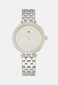 Reloj plateado con esfera redonda, adornado con cristales, acentos amarillos y una correa de pulsera metálica. Presenta manecillas doradas y logo.