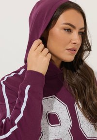 Mörkröd hoodie med vita ränder och en texturerad grafisk siffra 98. Har en dragsko i huvan och ribbade ärmslut för en bekväm passform.