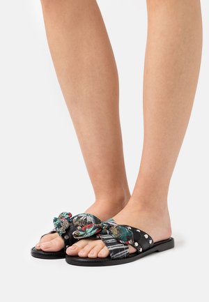 Sandales noires avec une bride à motif floral dotée d'un détail noué, ornées de clous en argent. Design ouvert à l'avant, semelle plate.