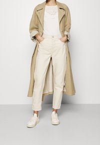 Beżowy trencz na białej bluzce z ażurowym zdobieniem, w połączeniu z jasnobeżowymi spodniami i białymi trampkami ze srebrnymi akcentami.