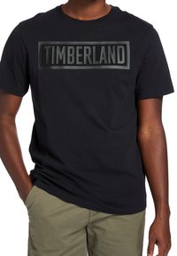 Camiseta de algodón negra de manga corta con un logo elevado en negro "TIMBERLAND" en un cuadro rectangular a lo largo del pecho. Diseño simple y clásico.