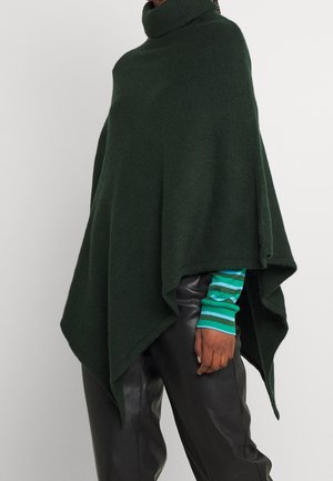Person trägt einen dunkelgrünen Rollkragenponcho über einem gestreiften grünen Langarmshirt und schwarzen Lederhosen, steht vor einem einfarbigen Hintergrund.