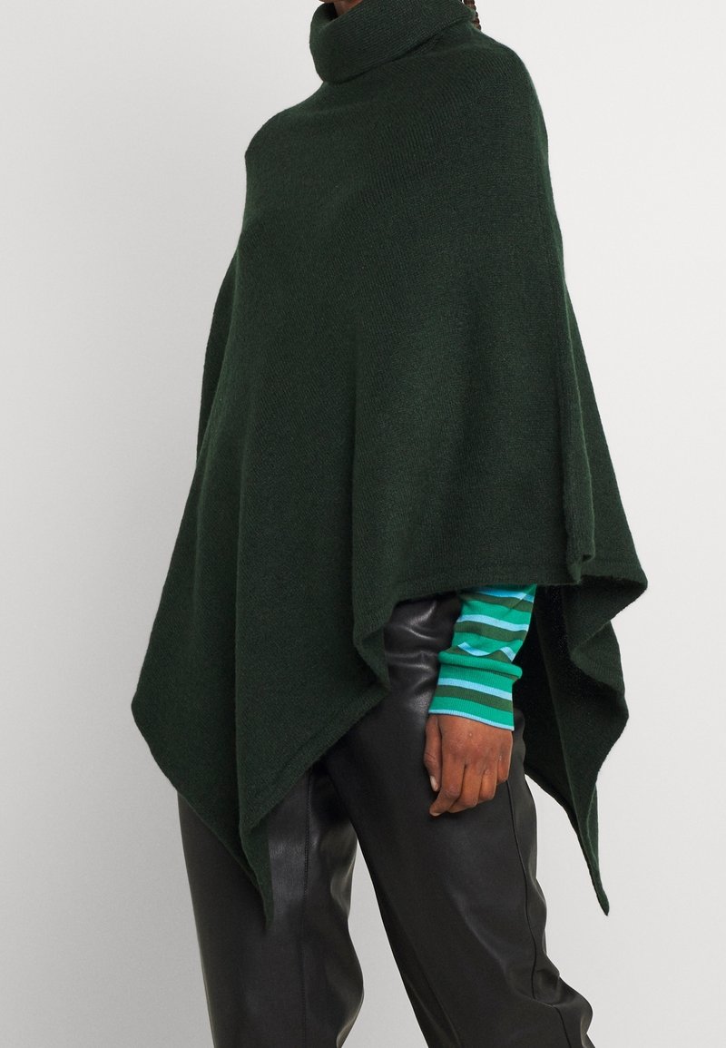 Personne portant un poncho à col roulé vert foncé sur une chemise verte rayée à manches longues et un pantalon en cuir noir, debout devant un fond uni.
