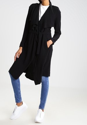 Femme portant un long manteau noir ceinturé, une chemise blanche, un jean skinny bleu et des baskets blanches, debout avec une main dans la poche.