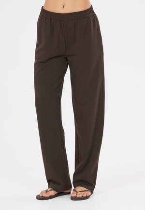 Personne portant un pantalon marron ample et des tongs noires, debout devant un fond blanc uni.