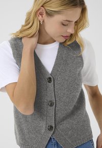 Grau gestrickte Weste mit V-Ausschnitt, ausgestattet mit vier dunklen Knöpfen, geripptem Saum und seitlichen Schlitzen, getragen über einem weißen T-Shirt und Jeans.