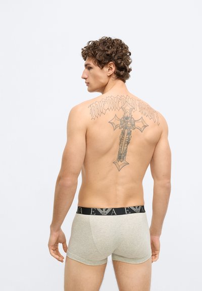 Beżowe bawełniane bokserki z czarną elastyczną gumą w pasie ozdobioną logo, noszone przez modela z widocznym dużym tatuażem na górnej części pleców.