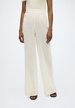 Kobieta nosząca kremowe, wysokie spodnie szydełkowane o szerokich nogawkach, czarne buty na obcasie z odkrytymi palcami oraz beżowy, prążkowany top z krótkim rękawem.