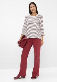 Gestreiftes Langarmshirt in Weiß und Maroon, kombiniert mit maronfarbenen, ausgestellten Hosen und passenden Sneakers. Hält eine maronfarbene Handtasche.