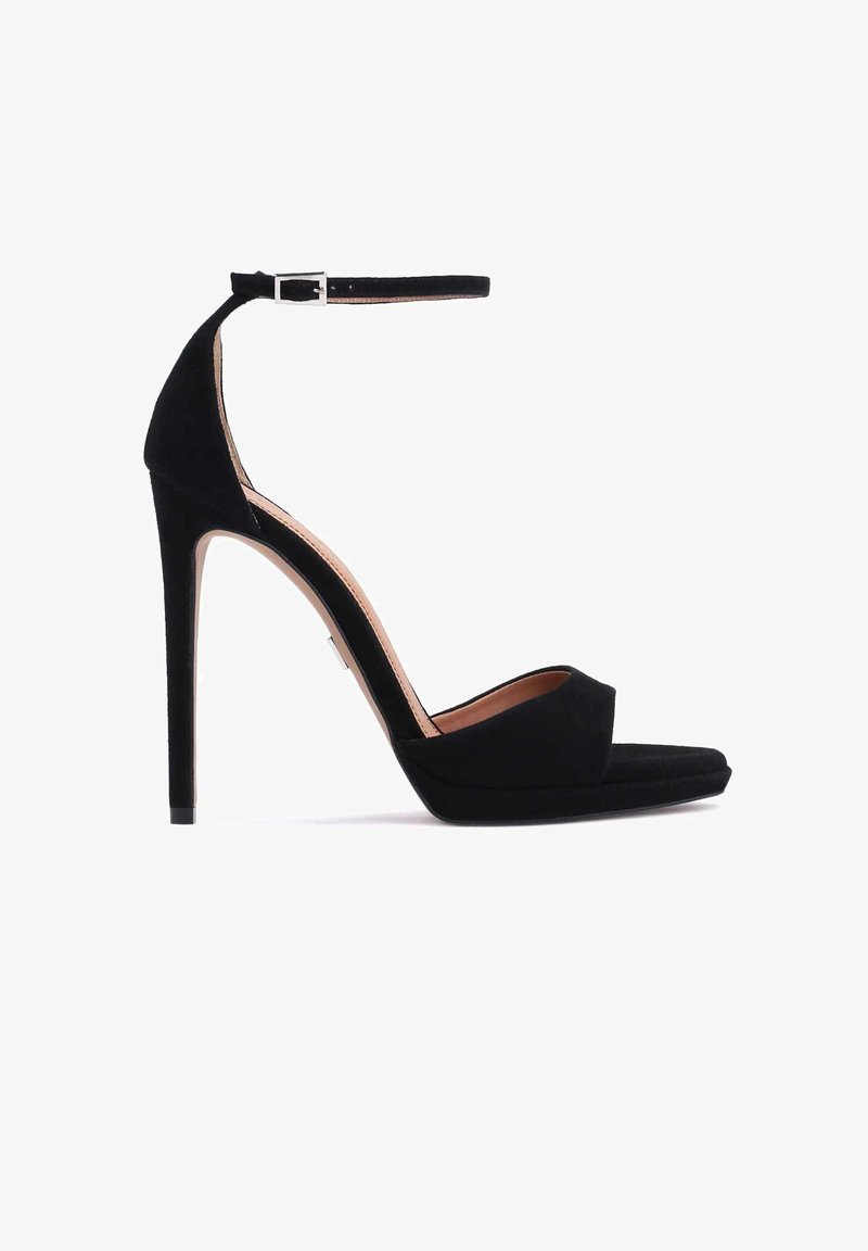 Sandale à talons hauts en daim noir avec bride à la cheville et boucle argentée, bout ouvert, et talon aiguille fin sur fond blanc.