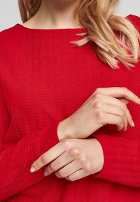 Red, textured sweater with a wide neckline, featuring long sleeves and a ribbed pattern. Hands crossed at the chest, displaying natural nail shape.