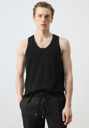 Jeune homme aux cheveux courts portant un débardeur noir sans manches et un pantalon noir à cordon, debout devant un fond clair uni.