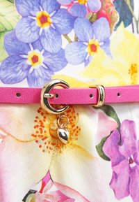 Ceinture en cuir synthétique rose avec une boucle circulaire dorée et un pendentif. Le fond présente un motif floral aux couleurs vives.