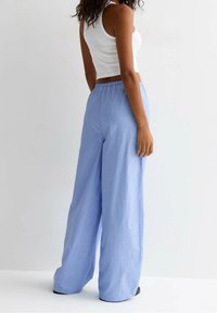 Vrouw in een witte mouwloze crop top en losse lichtblauwe gestreepte wijde broek, staande tegen een effen witte achtergrond.