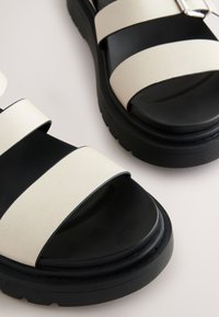 Witte leren sandalen met zwarte zolen, voorzien van twee brede banden en een gestructureerde afwerking. Accenten zijn onder andere een zilveren gesp op een van de banden.