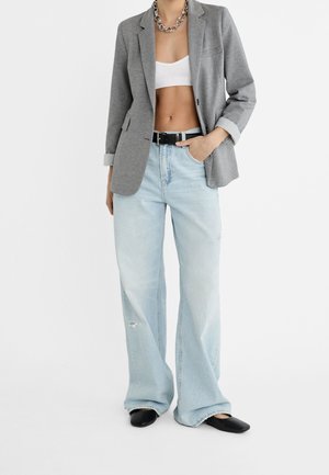 Blazer gris, top corto blanco, vaqueros de pierna ancha azul claro con dobladillo deshilachado, cinturón negro y zapatos planos negros; collar de cadena añade detalle.