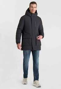 Zwarte donzen jas met capuchon, ritssluiting aan de voorkant, twee grote voorzakken en elastische manchetten. Gedragen over een blauwe spijkerbroek en lichte sneakers.