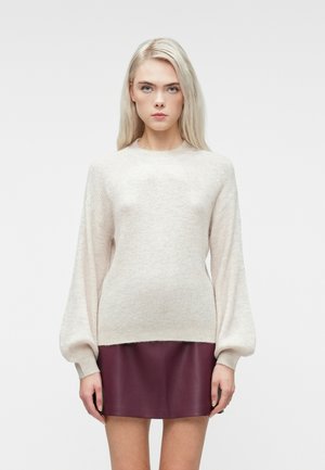 Femme aux longs cheveux blonds portant un pull en tricot beige clair avec des manches bouffantes et une jupe en cuir bourgogne courte, se tenant contre un fond blanc.
