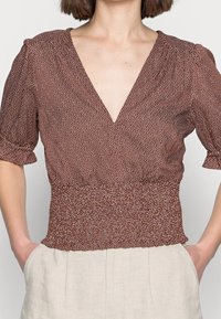 Brun mönstrad blus med djup V-hals, korta puffärmar och figursydd midja. Tillverkad av lätt material med vita prickar.