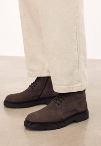 Botas de ante marrón hasta el tobillo con diseño de cordones, suela de goma y detalles de costura, combinadas con pantalones beige claros.