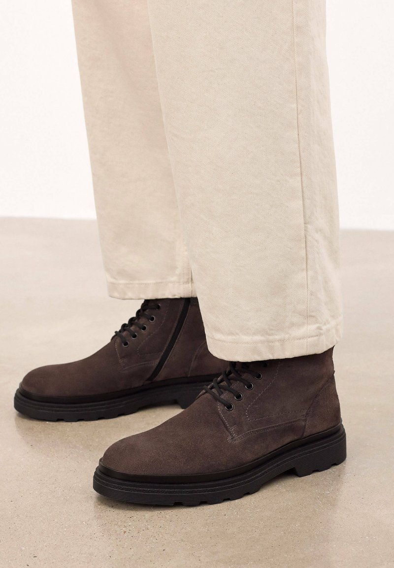 Botas de ante marrón hasta el tobillo con diseño de cordones, suela de goma y detalles de costura, combinadas con pantalones beige claros.