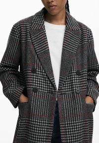 Abrigo de lana de cuadro de espiga gris, con cierre de doble botonadura en negro y detalles en rojo, que presenta solapas con muesca y bolsillos laterales, fabricado en tejido texturizado.