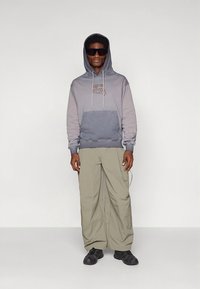 Modelis dėvi pilkos ir anglies spalvos džemperį su kanguros kišene, derinant su laisvomis šviesiai žaliomis kelnėmis ir juodais sportbačiais.