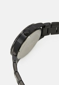 Čierne metalové hodinky s okrúhlym puzdrom, textúrovanou korunou a náramkom na články. Disponujú hladkým, matným povrchom a jednoduchým, nezdobeným zadným dielom.