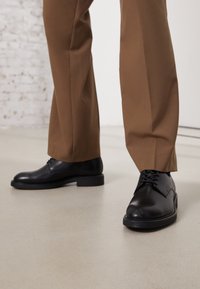 Scarpe in pelle nera con superficie liscia e punta rotonda, caratterizzate da chiusura con lacci e suola bassa e piatta. Indossate con pantaloni marroni.
