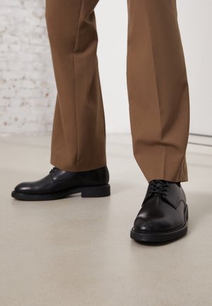 Black leather shoes with smooth surface and round toe, featuring lace-up closure and a low, flat outsole. Paired with brown pants.