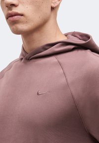 Κοντινό πλάνο ενός ατόμου που φοράει μωβ φούτερ Nike με εμφανές λογότυπο Nike, ανοιχτόχρωμο δέρμα με ακμή και ένα μικρό κρίκο στο αριστερό αυτί.