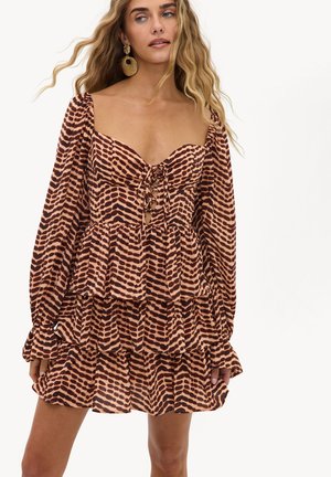 Femme aux longs cheveux ondulés portant de grandes boucles d’oreilles en or et une robe marron à volants avec de longues manches bouffantes et un laçage à l’avant.