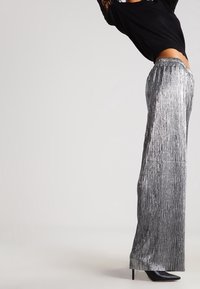 Osoba oblečená v lesklých stříbrných širokých kalhotách s plisováním, černých lodičkách na vysokém podpatku a černém topu s krajkovými rukávy, s rukama vztyčenýma nad hlavou.