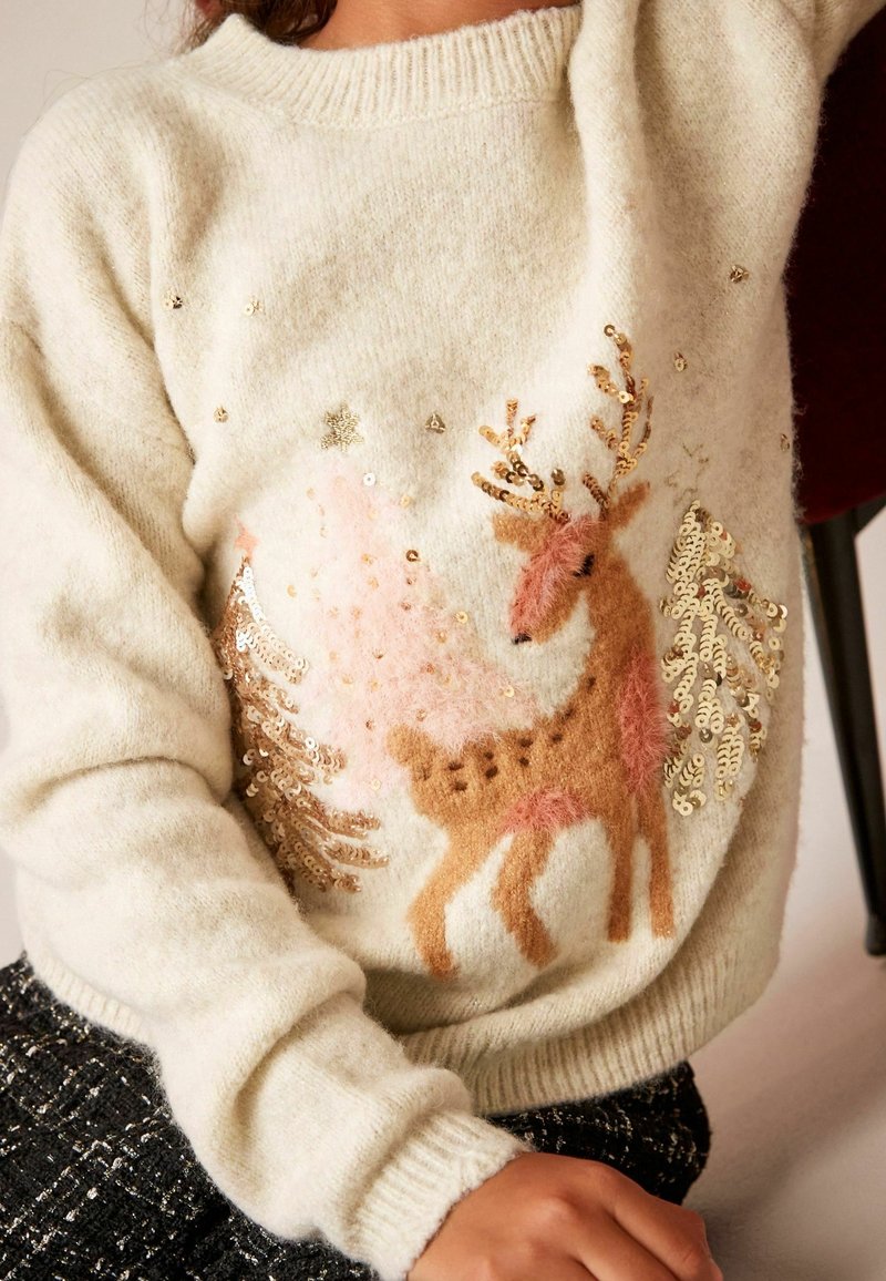 Pull crème douillet avec un renne brun à fourrure texturée et des arbres en sequins dorés, porté par une personne avec un bras levé.