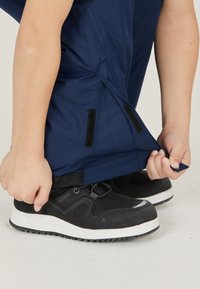 Marineblauwe waterdichte broek met klittenbandsluitingen bij de enkels, die met de handen worden aangepast boven zwarte sneakers met witte zolen.
