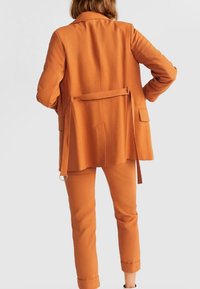 Oranžový blazer se stuhou na zádech, spárovaný s odpovídajícími kalhotami. Tkanina vypadá hladce, blazer má dvě zadní kapsy.