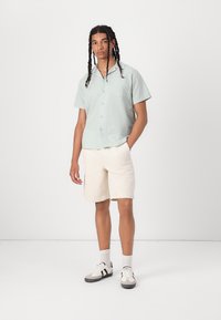 Lys grøn kortærmet skjorte med knaplukning i struktureret stof, parret med beige shorts. Modellen bærer hvide og grå sneakers og står på en neutral baggrund.